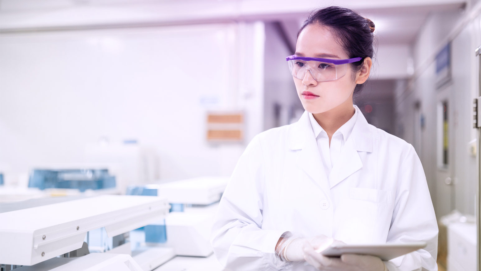 Scientist in a lab coat and protective eyewear holding a tablet in a modern laboratory, representing advanced scientific research.