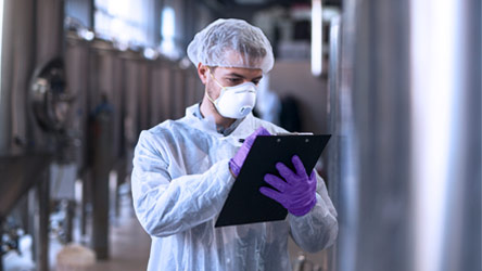Scientist in protective gear and mask inspecting equipment and taking notes in a lab, representing safety and precision.