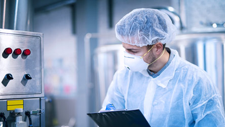 Scientist in protective gear and mask monitoring industrial equipment and taking notes, symbolizing safety and quality control in research.