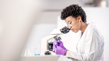Scientist in a lab coat and gloves examining samples under a microscope, representing detailed scientific research.