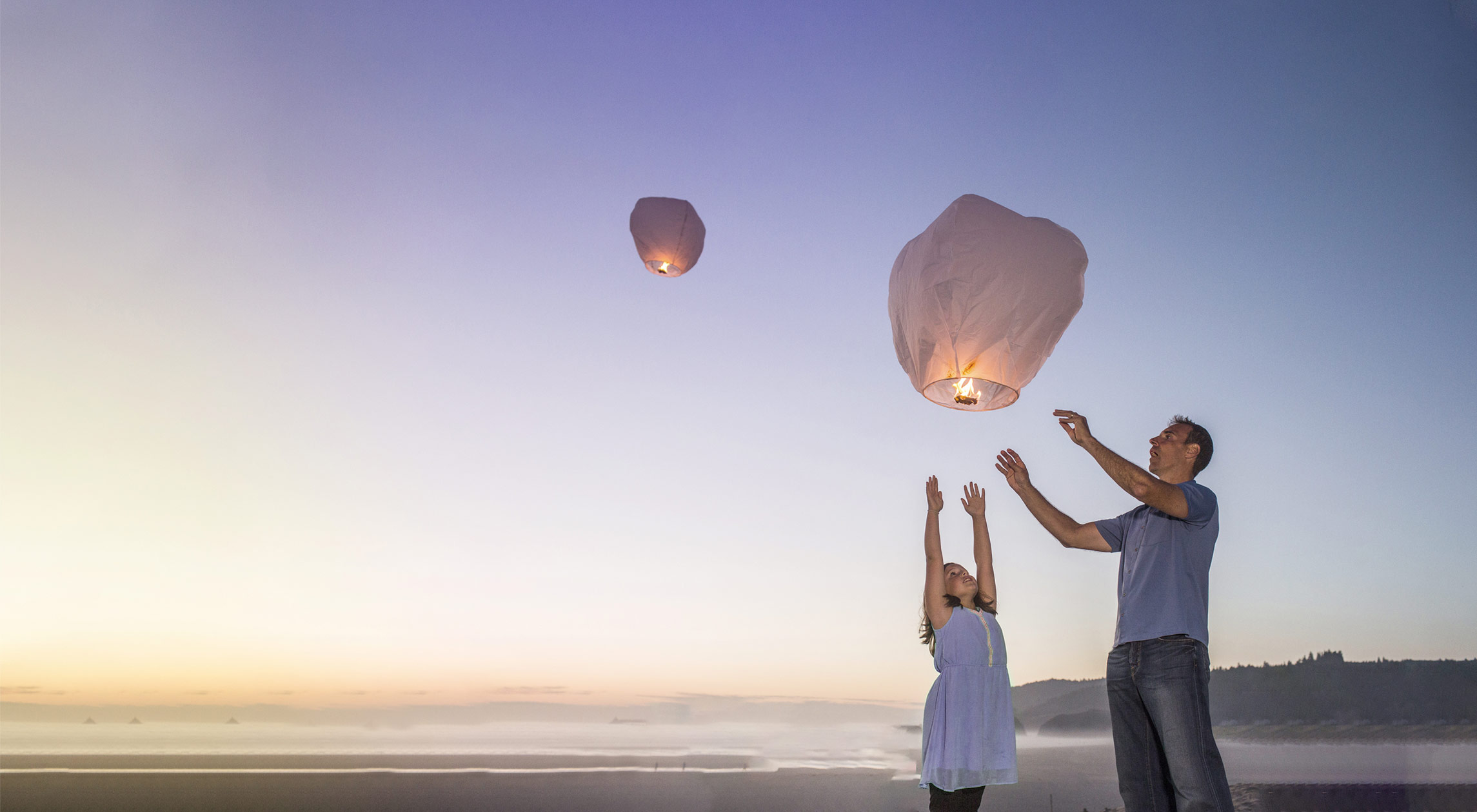 Father and daughter releasing sky lanterns at sunset on the beach, symbolising hope, dreams, and togetherness.