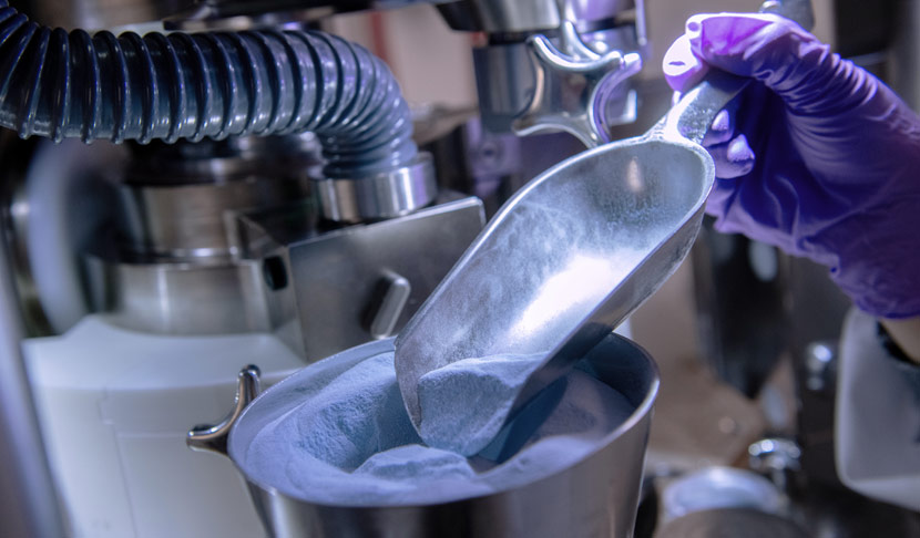 Scientist wearing purple gloves scooping a powdered substance in a lab, representing precision in pharmaceutical manufacturing.