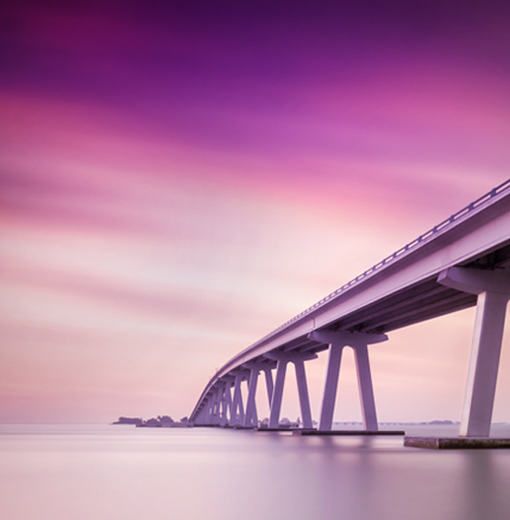 Long bridge extending over calm water during a purple and pink sunset, symbolising connection and tranquillity.