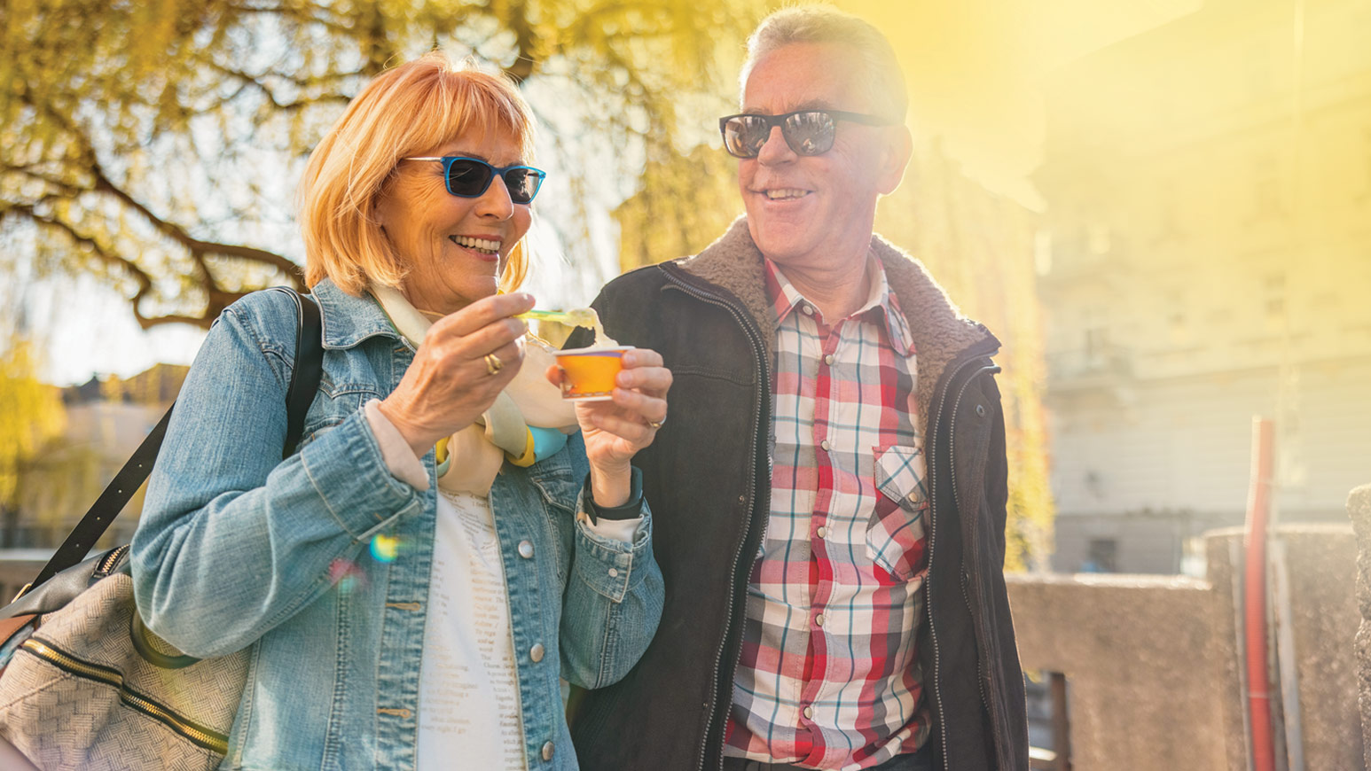 Smiling senior couple enjoying outdoor yogurt snack in sunny weather, both wearing sunglasses and casual clothing.