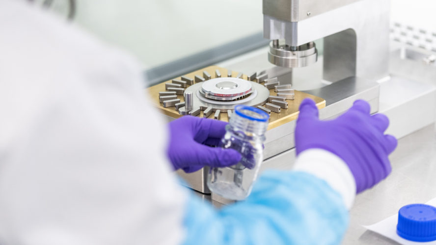 Lab technician wearing gloves operates equipment, handling a vial during a pharmaceutical manufacturing process.